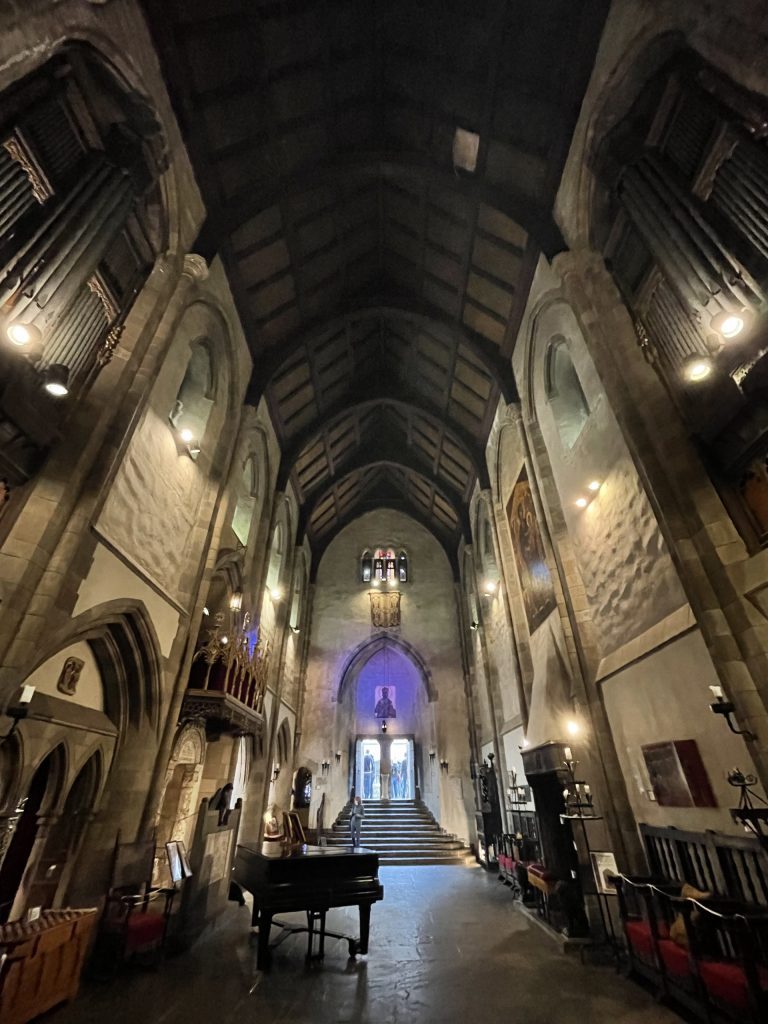 Photograph of the large dimly-lit hall of Hammond Castle, with a soaring wooden ceiling and stone walls, with a stained glass window at the back end, and a piano in the center.