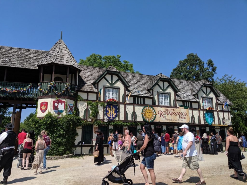 Entrance gate of the Bristol Renaissance Faire, a Tudor-style architectural construction of faux plaster and exposed timber beams, adorned with painted wooden heraldry and covered in climbing ivy.