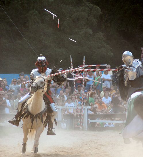 Two armored men on horseback run at each other with lowered lances, which break into wooden shards upon impact with their metal shields while a crowd of spectators observes in the background.