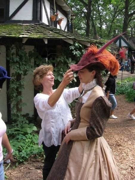 One woman dressed in a white blouse puts an elaborate, burgundy-colored feathered hat on another woman dressed in a Tudor-style gown.