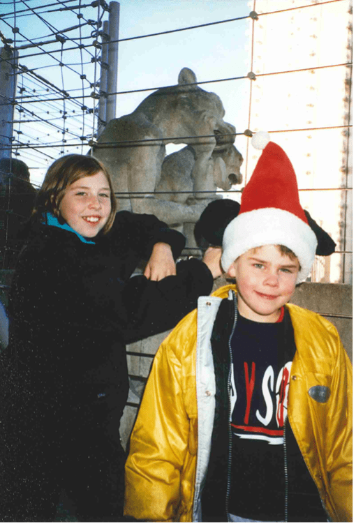 A girl and boy smiling and standing with a metal fence behind them. The boy wears a red fuzzy hat.