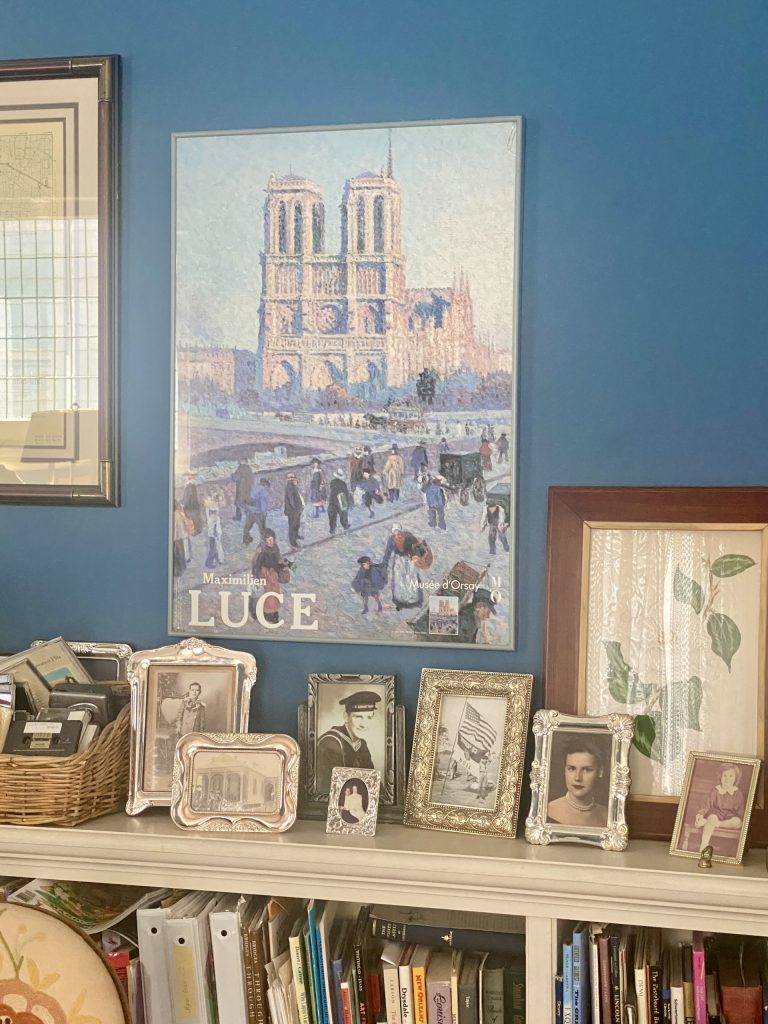 A photograph of a print of a painting hanging on a wall above a bookcase. There are numerous old framed photographs on the top of the bookshelf.
