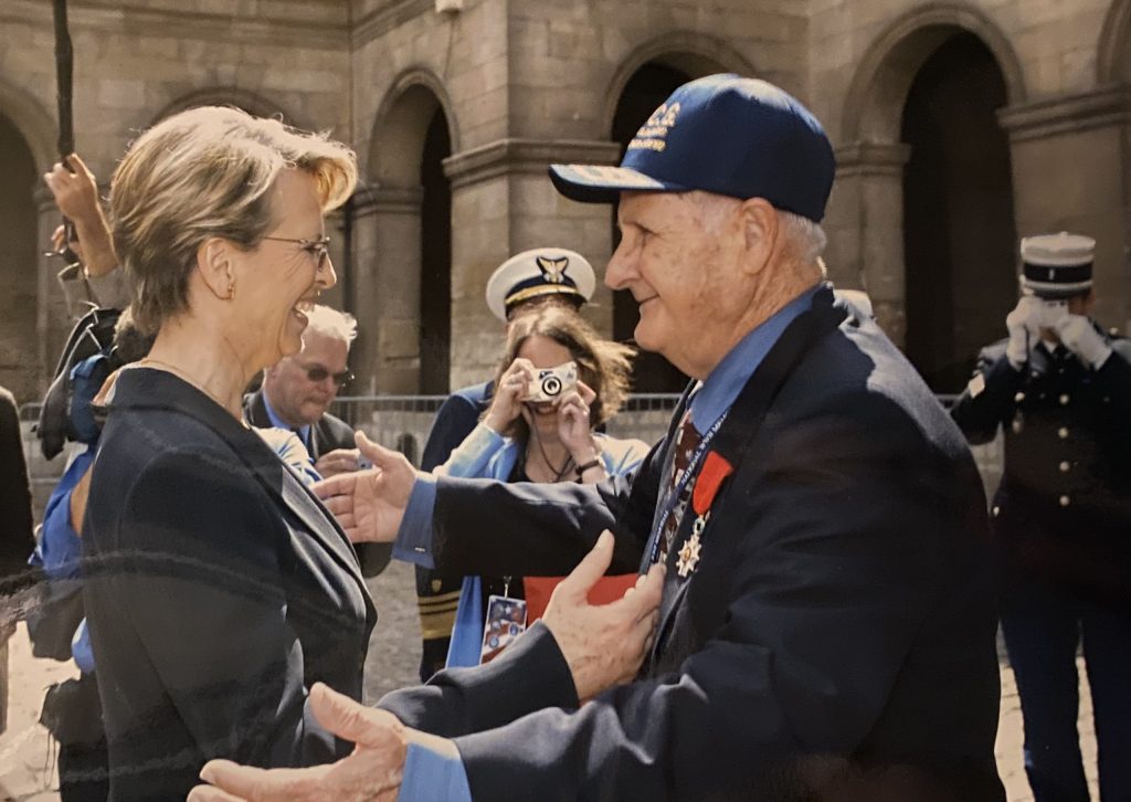 A woman and man greeting each other with open arms while people look on from behind them.