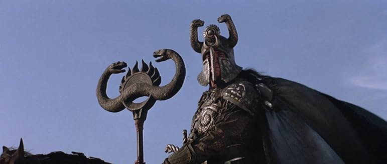 Figure 1: A still presenting the sorcerer Thulsa Doom wearing black scaled armor and a billowing cape against a blue sky. Beside him is a crest that depicts two conjoined snakes facing each other with a sun symbol mediating them.
