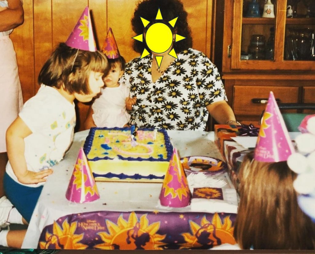 Color photograph of a birthday party with themed cakes, napkins, plates, hats, and presents on a table surrounded by party attendees.