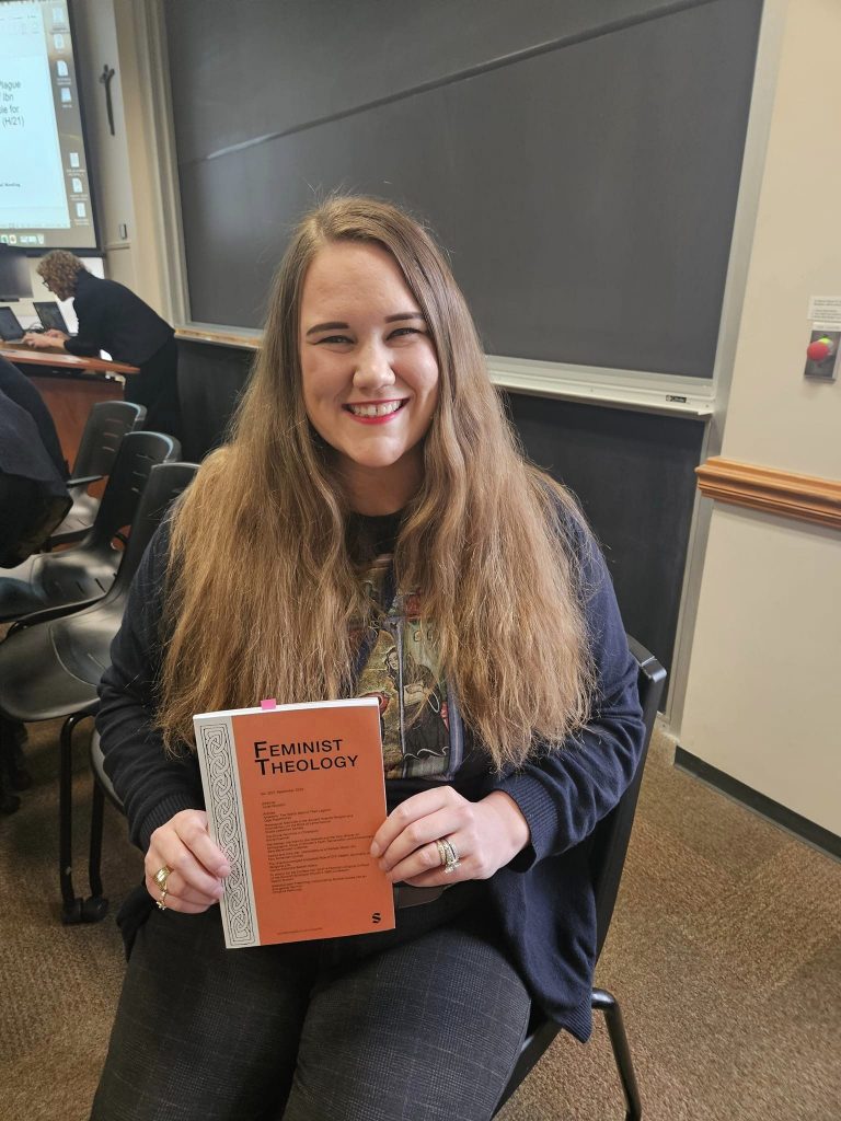 Person smiling while holding a book titled "Feminist Theology" in a classroom.