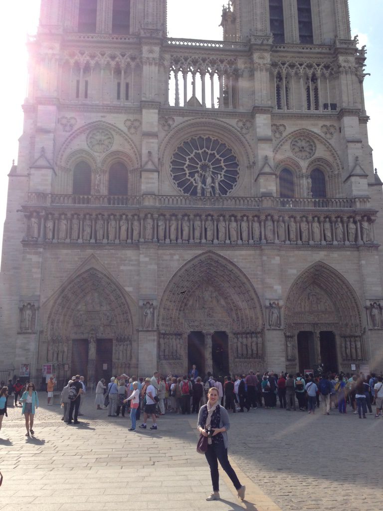 Color photograph of a woman holding a camera in front of Notre Dame with crowd behind her.