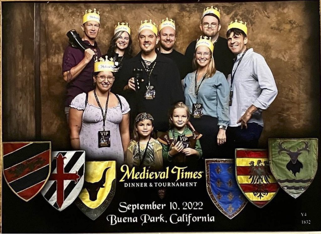A cheerful group of eight adults and two children at Medieval Times Dinner and Tournament; several wear paper crowns and medievalizing attire.