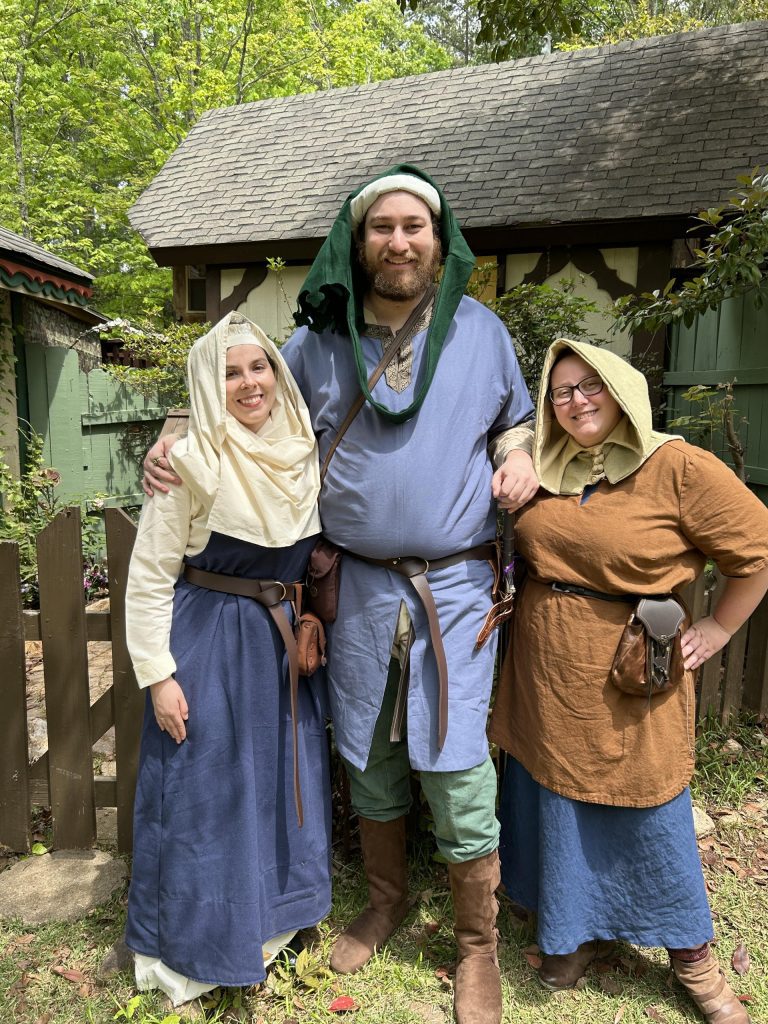 Three people dressed in pseudo-historical dress posing in front of a cottage at the Georgia Renaissance Festival. One wears a wool dress and wimple, one wears a liripipe and tunic, and one wears a hood with a tunic over a dress.