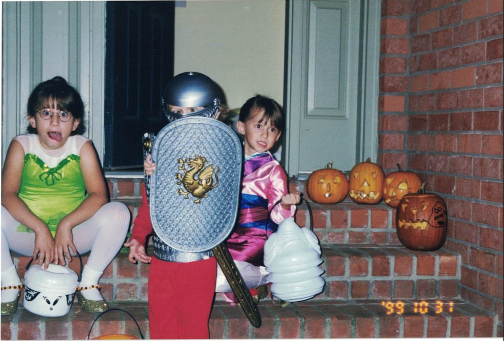 Three children pose on brick steps in Halloween costumes (Tinkerbell from Disney’s Peter Pan, a medieval knight, and Mulan from the Disney film). The children are making funny faces, holding Halloween themed buckets, and sitting beside four carved pumpkins.