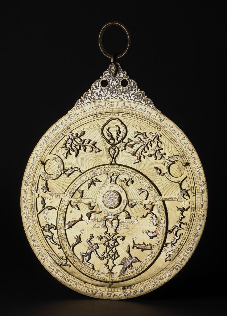 An astrolabe inscribed in Arabic, featuring delicate colored metalwork decorations of plants, animals, and a man along the inner and outer rings.