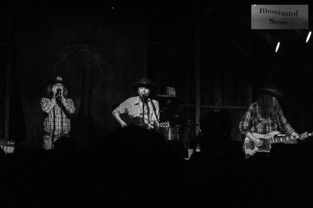 Colter Wall, a folk musician, sits on stage in Codfish Hollow, Maquoketa, Iowa. He plays guitar and is accompanied by a man playing bass guitar (right) and a man playing harmonica (left).