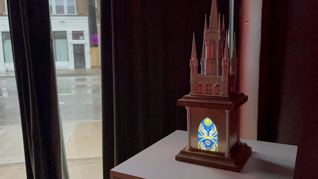 A small replica of a medieval church with tall spiky towers and an illuminated colorful window resting on a table beside a window.