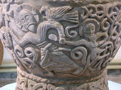 ALT: Detail of a stone baptismal font showing Christ with a halo and cross pulling another figure with a halo.