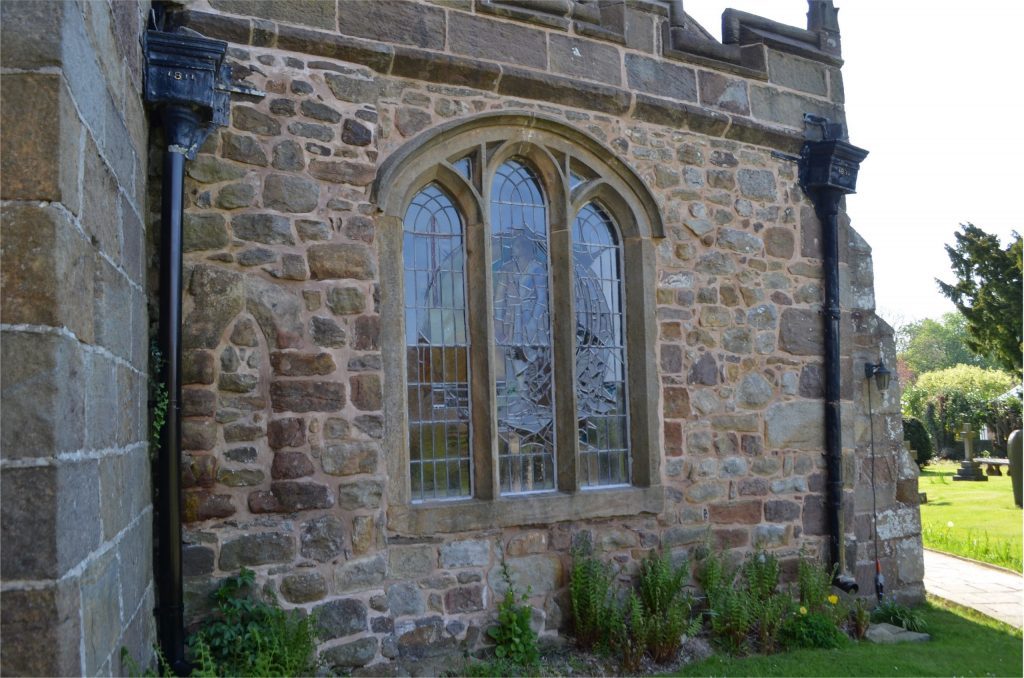 South aisle of stone church building from the exterior showing small, filled in lancet window, and larger, three light window.