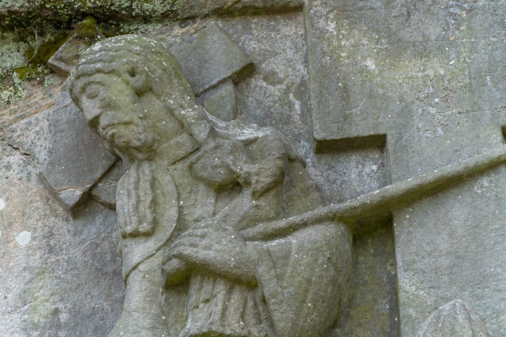 Long-haired man with a cross behind his head holds a kneeling headless figure on his left arm.