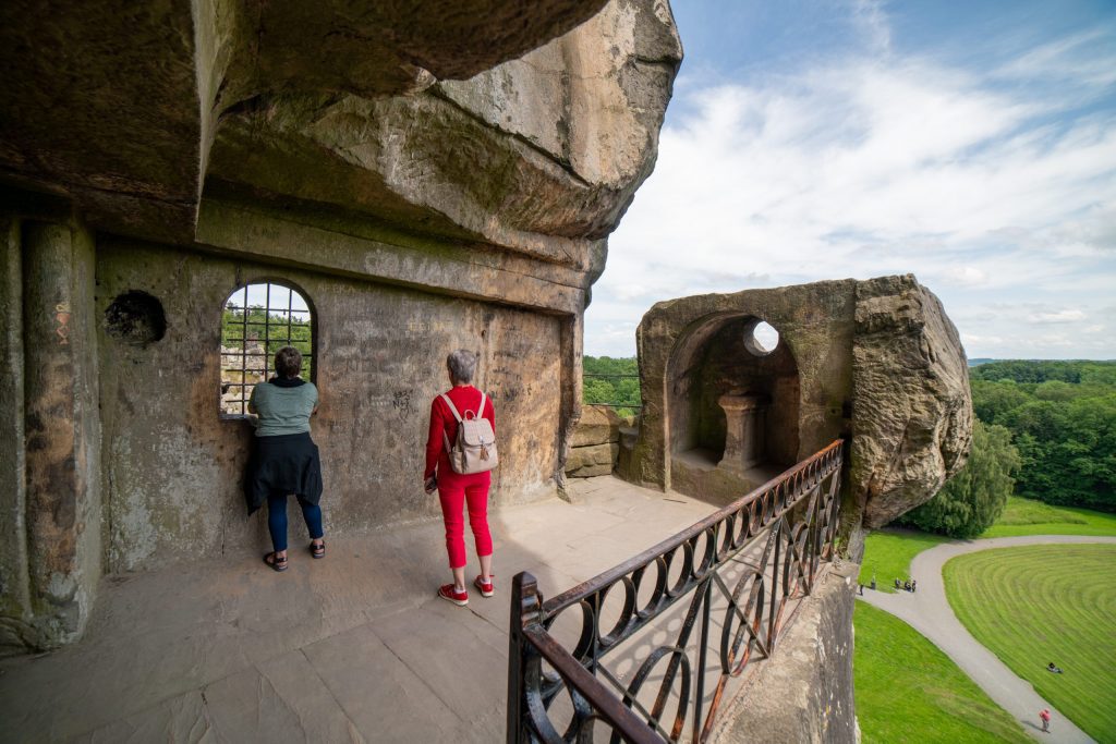 Two people face a window cut into a rock wall. The other walls and ceiling are missing and so overlook a grassy field below.