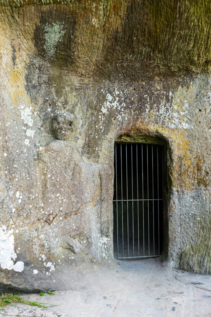 Shallow relief carving of a standing and gesturing man next to a barred entrance to a cave.