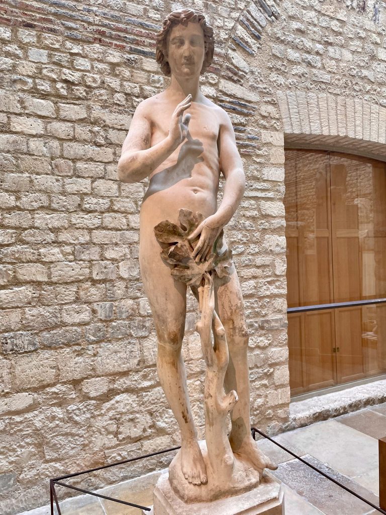 A stone statue of a naked man with a tree between his legs. He holds a large leaf over his crotch.