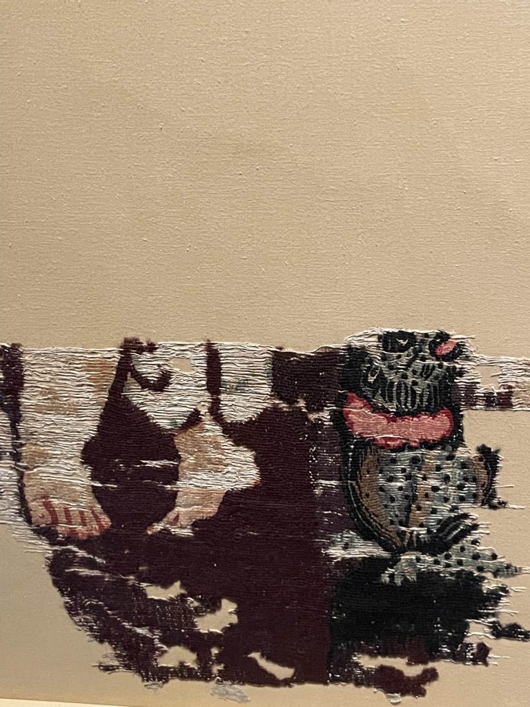 A very work textile fragment that depicts two feet and a vessel of some sort. The background is black.