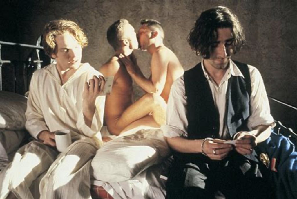 Screenshot from Derek Jarman's Edward II showing four people on a bed, the two in the background are unclothed while the other two read letters.