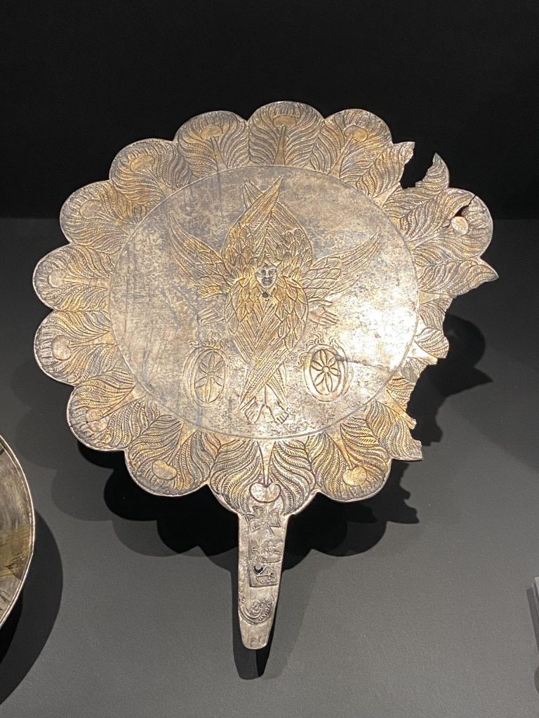 A close up of an old metal mirror with decorative incisions on it, including an image of a seraphim in the center.