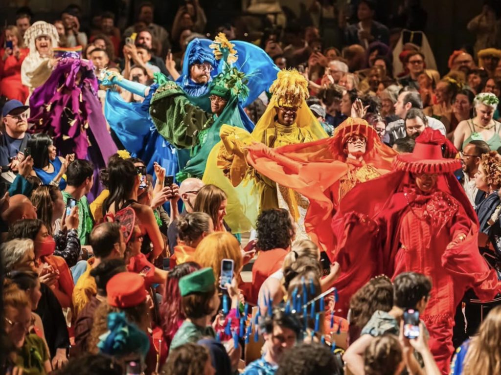 A procession of people in rainbow garments surrounded by a crowd of people -- some applaud, some take photos on their phones.