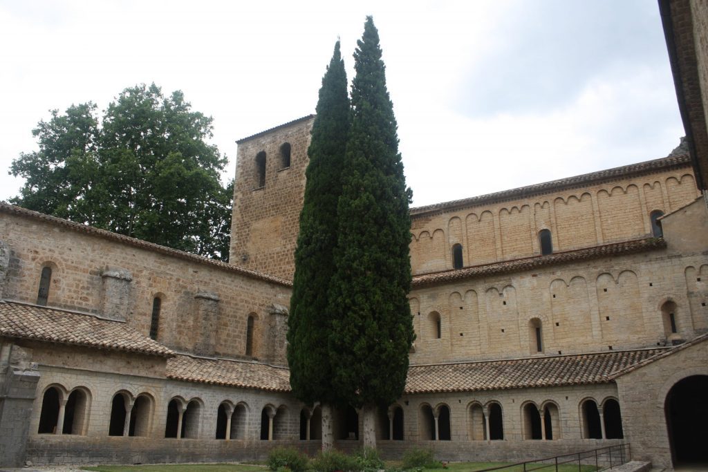 An open courtyard is surrounded on two sides by a covered walkway and stone buildings. Two tall cypress trees grow in the courtyard’s corner.