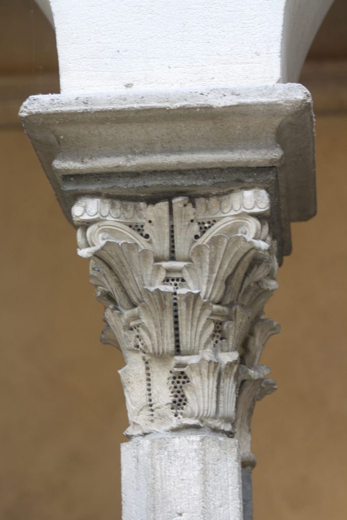 A stone capital decorated with three tiers of upright leaves that curl forward slightly, interspersed with a dense grid of drill holes.