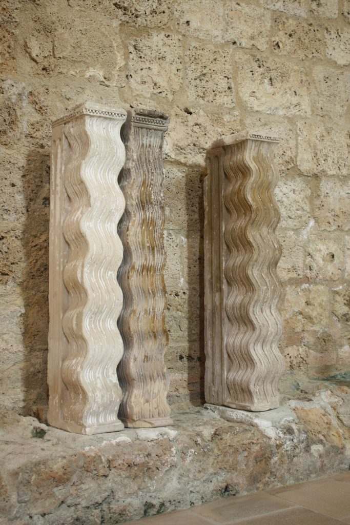 Three stone columns carved to resemble rows of waves lean against a stone wall.
