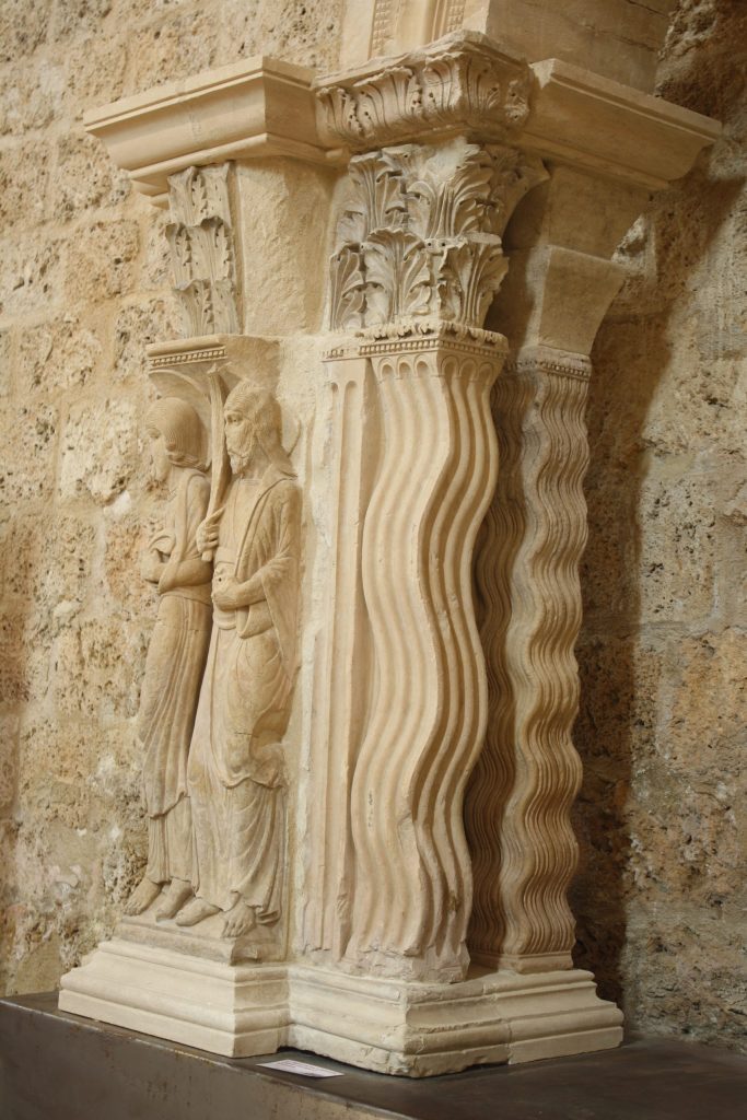 A pair of stone columns carved to resemble rows of waves support capitals and horizontal slabs decorated with leaves.