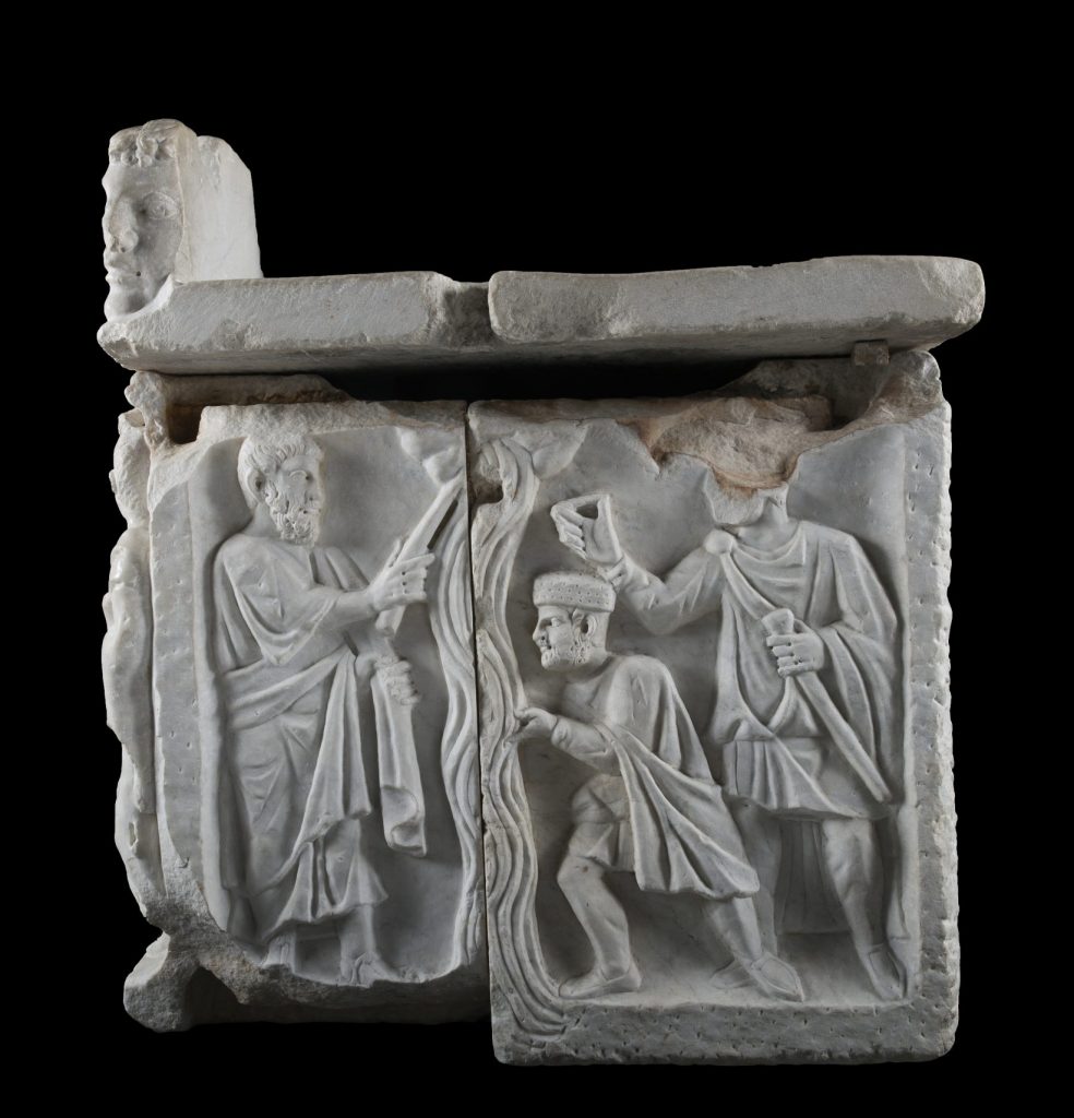 A stone relief sculpture shows three men surrounding a wavy horizontal column; the man at left strikes the top of the column with a stick.