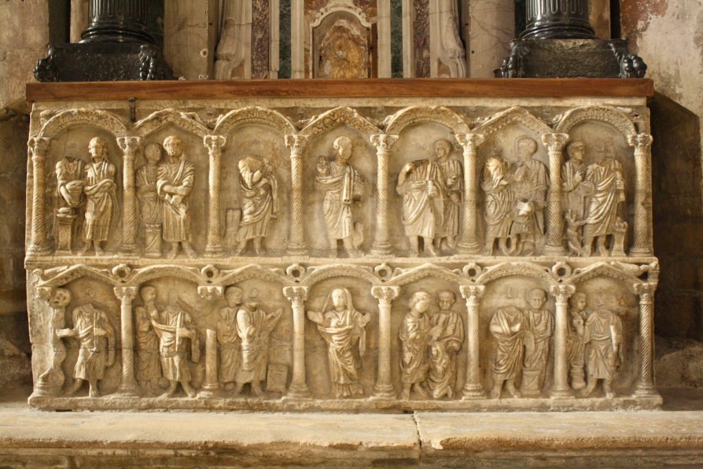 A stone relief sculpture showing two tiers of seven figure groupings, each grouping framed by arches and columns.