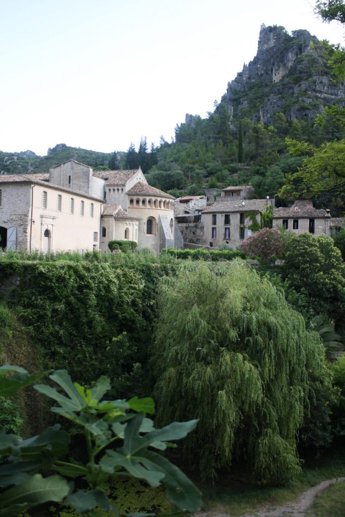 A church sits at the edge of a steep ledge covered in plants. A tall cliff towers overhead.
