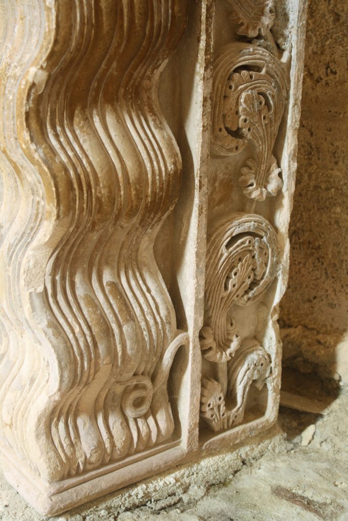 A close-up look of a column carved to resemble rows of waves showing a small carved snake next to a row of plants.