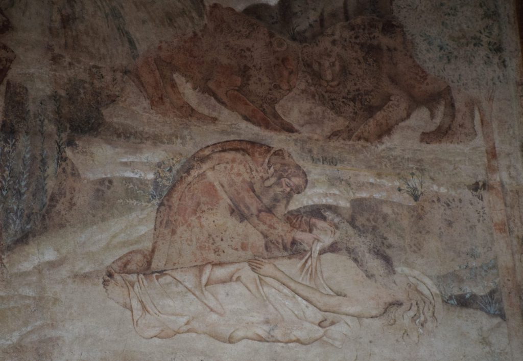 Part of a wall painting showing one monk leaning over the dead body of an elderly man, lying on a ledge of flat rocks.