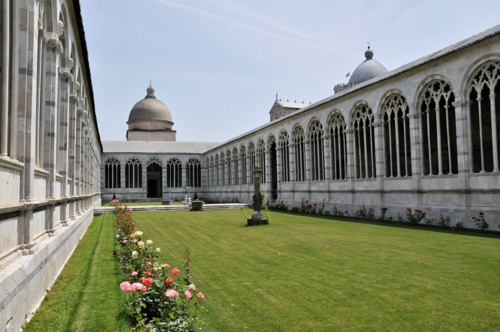 View of the Camposanto cemetery, Pisa (Italy), showing central burial ground and surrounding galleries. The dome of Pisa Cathedral is just visible on the right, beyond the Camposanto.