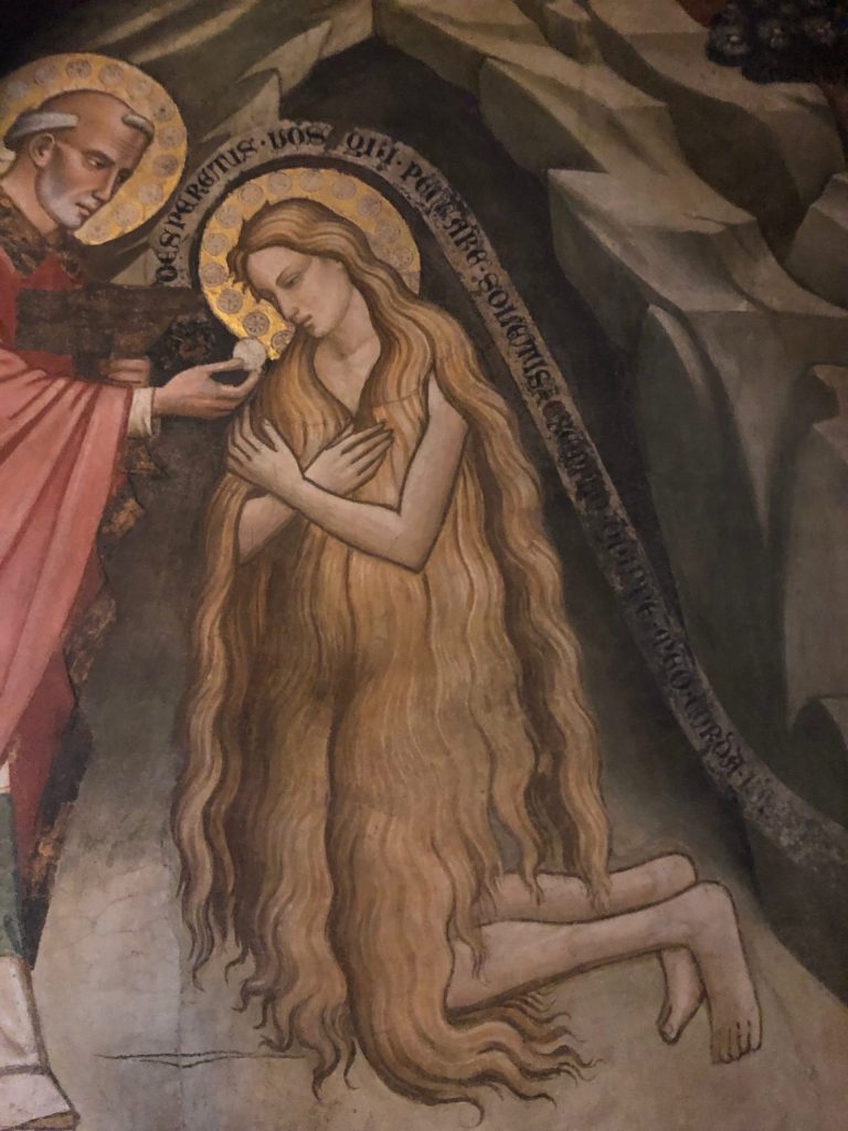 Detail of a fresco with Mary Magdalen, clad only in her hair, kneeling at the mouth of her cave. Facing to the left she receives Last Communion from St. Maximin. A scroll wraps over her halo and follows the outline of her body reading: NE DESPERETIS VOS QUI PECCARE SOLETIS EXEMPLOQUE MEO VOS REPARATE DEO or “do not despair those of you who are accustomed to sin, and in keeping with my example, return yourselves to God.”