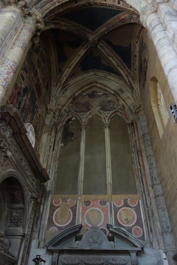 Alt Text: A view looking up into a dark, shallow and narrow chapel in a state of some disrepair. High on the walls, just under the vaulting are three frescoes depicting the life of the Magdalen: two on the left, one on the right.