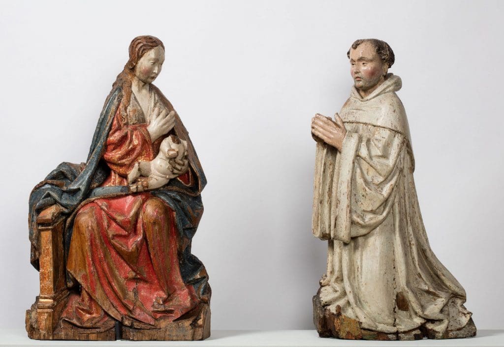 Two wooden sculptures, the one on the left representing Mary, seated with the (now decapitated) Christ child on her lap, squeezing her breast; on the right St Bernard kneeling in prayer.