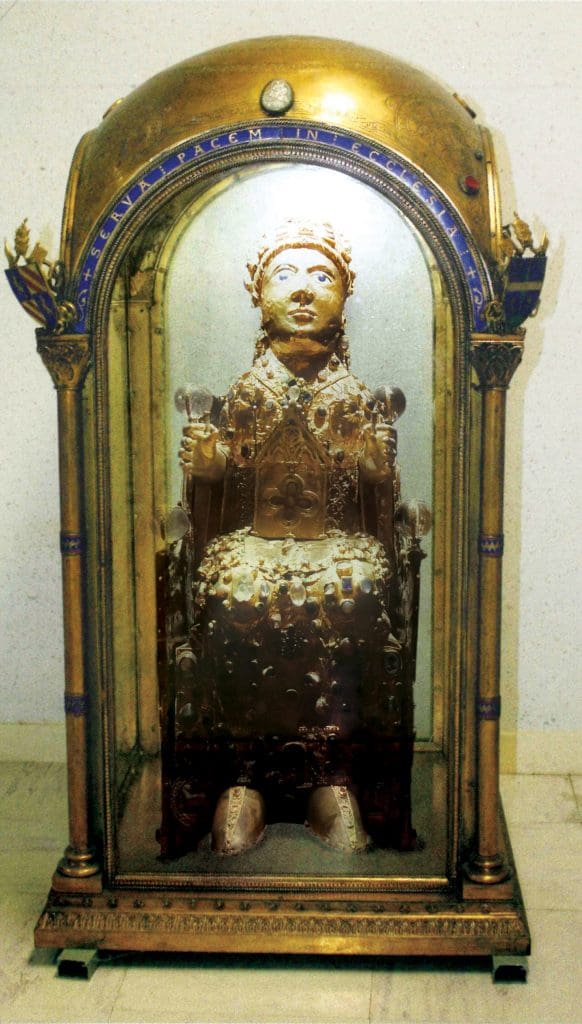 Color photograph of a small sculpture of a seated girl encrusted with gems located within a golden shrine with clear glass panel sides.