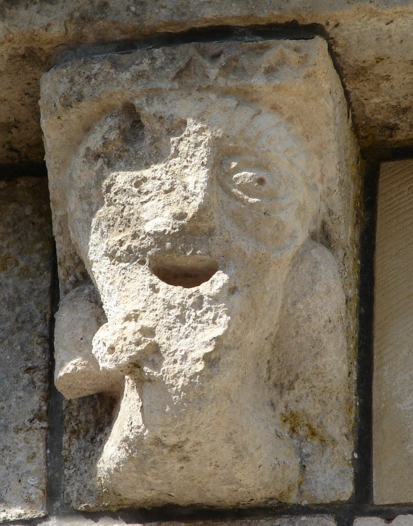 Leering corbel figure stroking his beard while his large eyes look to his left.