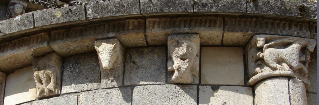 Leering corbel figure in situ looking at a capital next to him, featuring two large beasts engaged in sodomy.