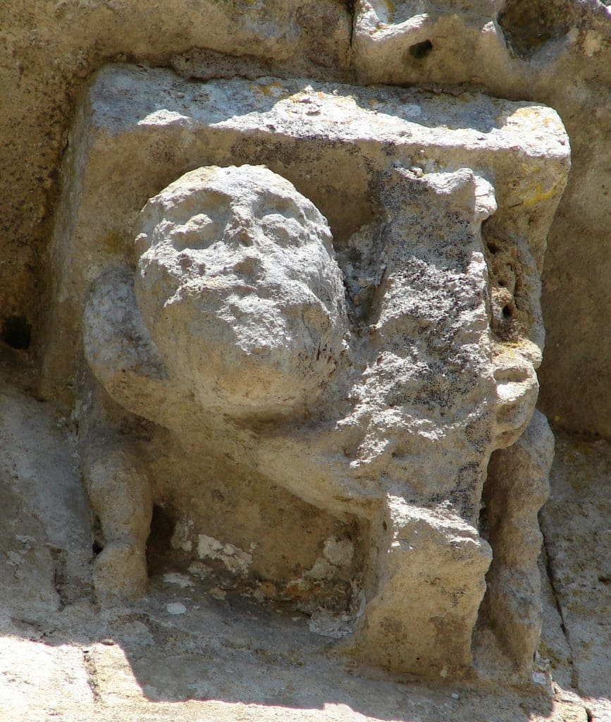 A corbel figure crouches with legs very far apart as he plays a harp.