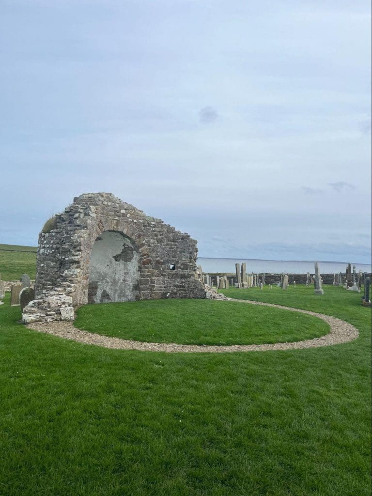A circular ruined church, with the sea and a graveyard visible in the background.