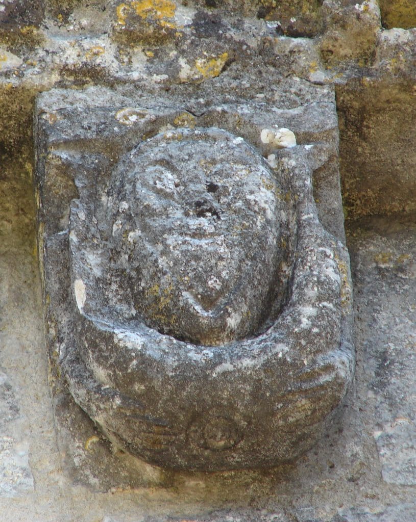 Corbel figure with feet around ears, exposing anus.