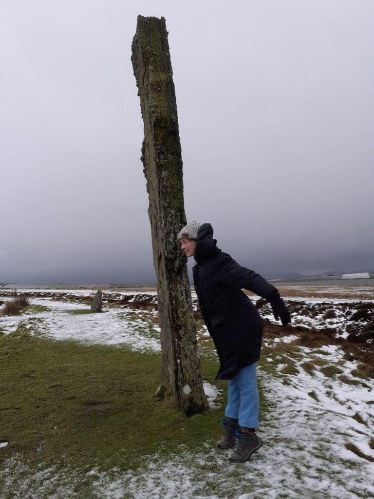 A female person wearing a winter coat leaning forward next to a standing stone on a snowy day.