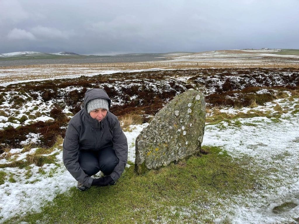A female person crouching next to a standing stone on a snowy day.