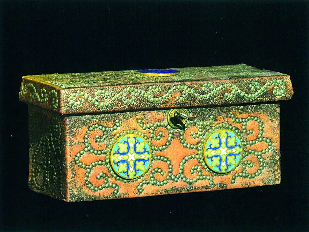A color photograph of a small rectangular metal box with colorful, enamel decorations made up of swirling lines of dots and circles with cross patterns inside of them.