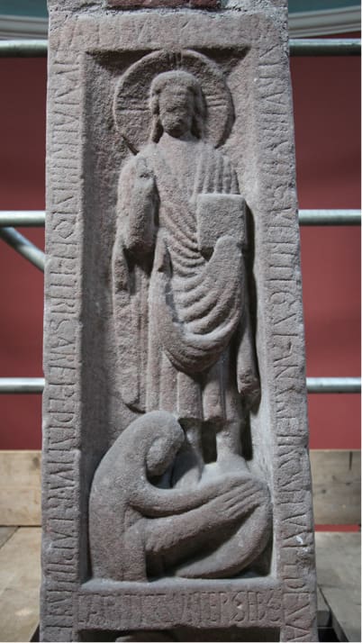 A close-up of the panel of the Ruthwell Cross that depicts Mary Magdalene washing Christ’s feet. Both figures are carved in relief on pink-grey sandstone. Latin inscriptions in Roman capitals are engraved in all four borders.